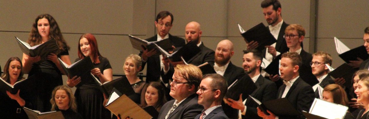 Choir in formal black attire singing onstage, holding open music folders during a performance.
