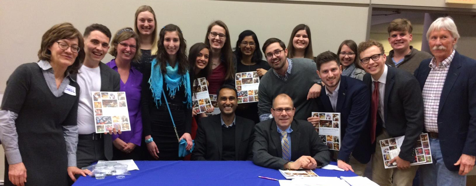 Group of adults posing behind a table at an indoor event, with several people holding printed photo-collage sheets.