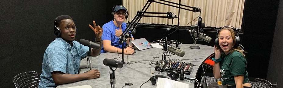 Three people wearing headphones in a podcast studio sit around a table with microphones and audio equipment, smiling and gesturing toward the camera.