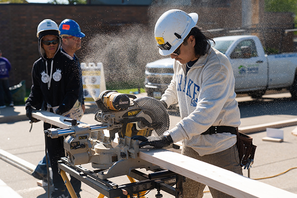 Drake Student in a hard hat and safety glasses uses a miter saw to cut lumber outdoors, with sawdust in the air and other workers in the background.
