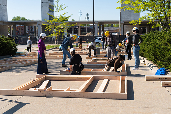 Group of volunteers wearing hard hats assemble wooden frame sections on a paved outdoor site near a large concrete building.