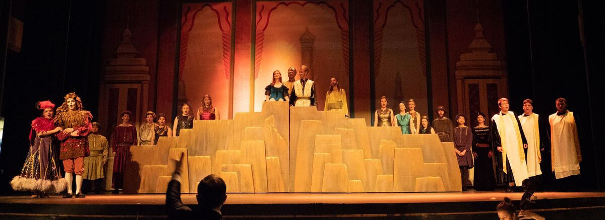 Cast of a stage musical standing on a large stepped set under warm theatrical lighting, framed by an ornate palace backdrop.