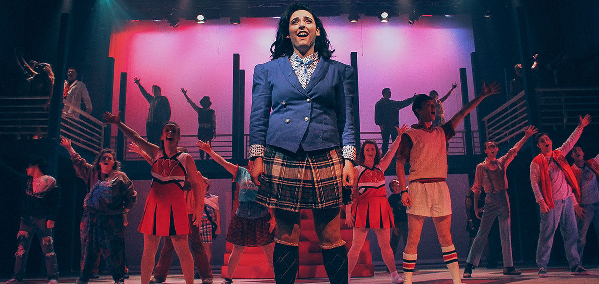 Stage musical performance with a central actor in a blue blazer and plaid skirt, surrounded by an ensemble with raised arms under colorful pink and purple lighting.