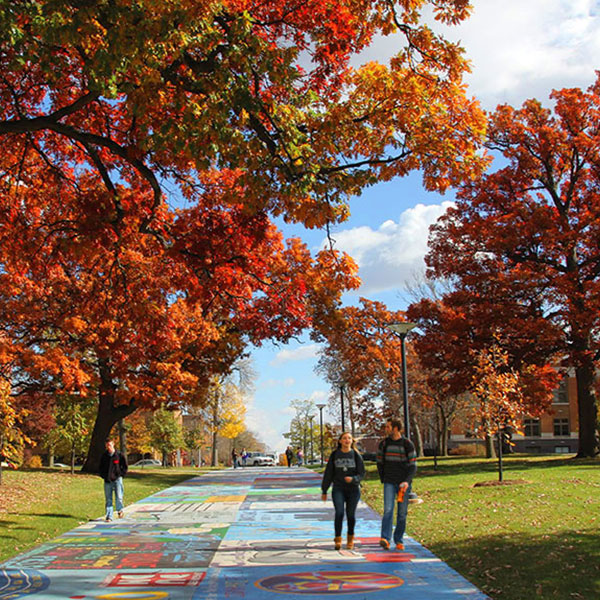 Campus walkway painted with colorful murals, lined with red and orange autumn trees as students walk past brick buildings under a partly cloudy sky.