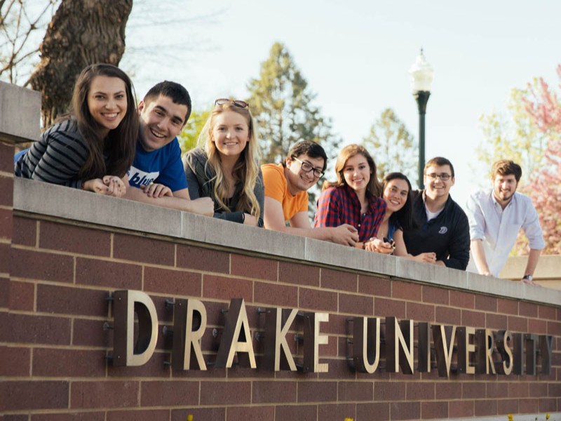 A group of eight students lean on the brick ledge about the Drake University sign