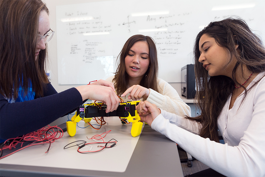 For a student competitive club, three Drake students collaborate on a walking robot in a classroom with a whiteboard in the background