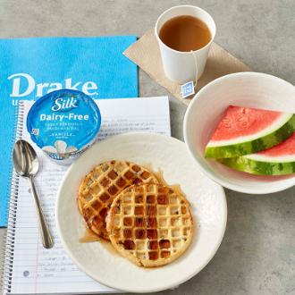 A cup of coffee next to two plates of waffles and watermelon sitting on top of a notebook on a table inside of a Drake University dining hall