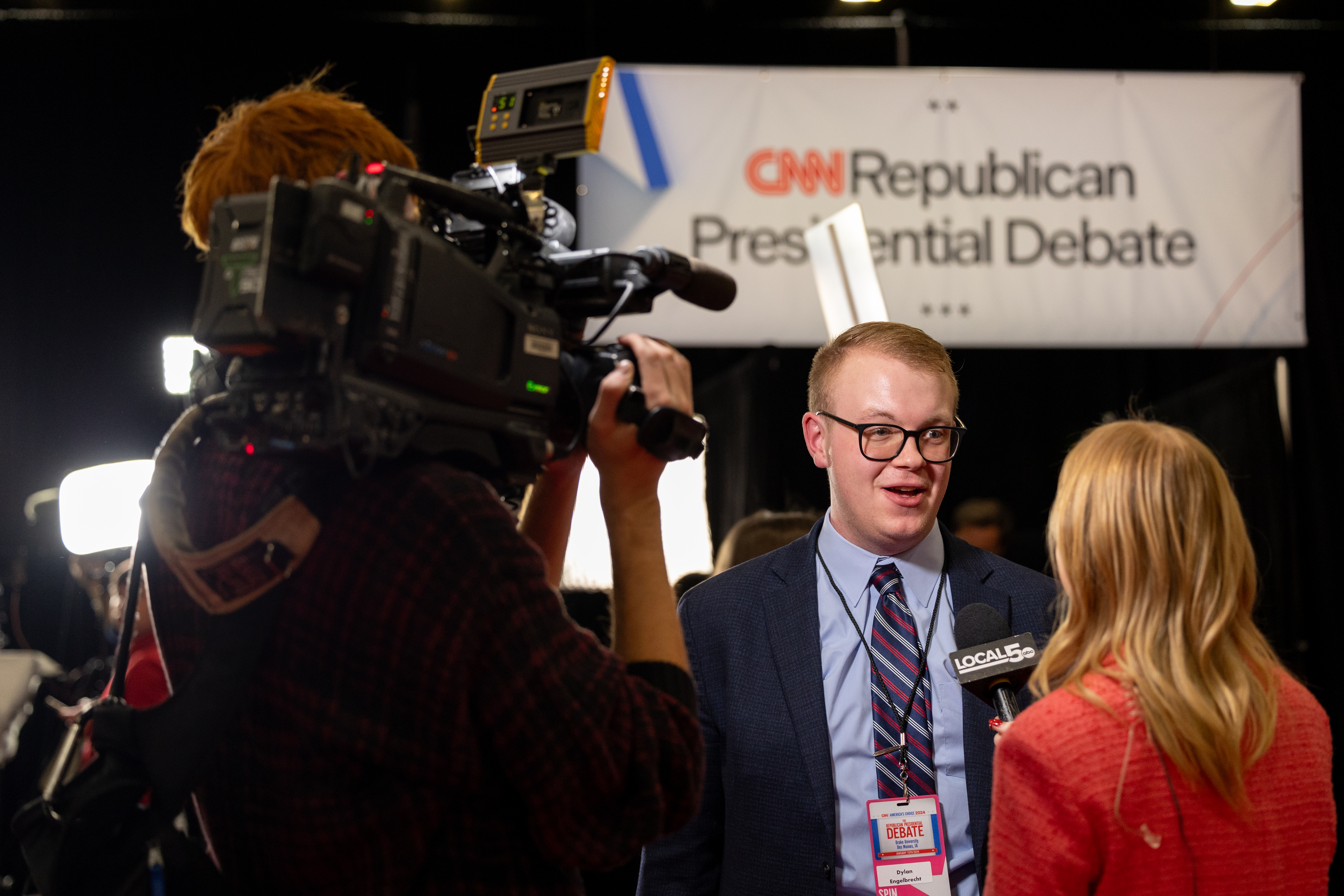 A camera focuses on a local newscaster interviewing a Drake student, in a suit and with a press pass, behind the scenes at the Iowa Republican Presidential Debate