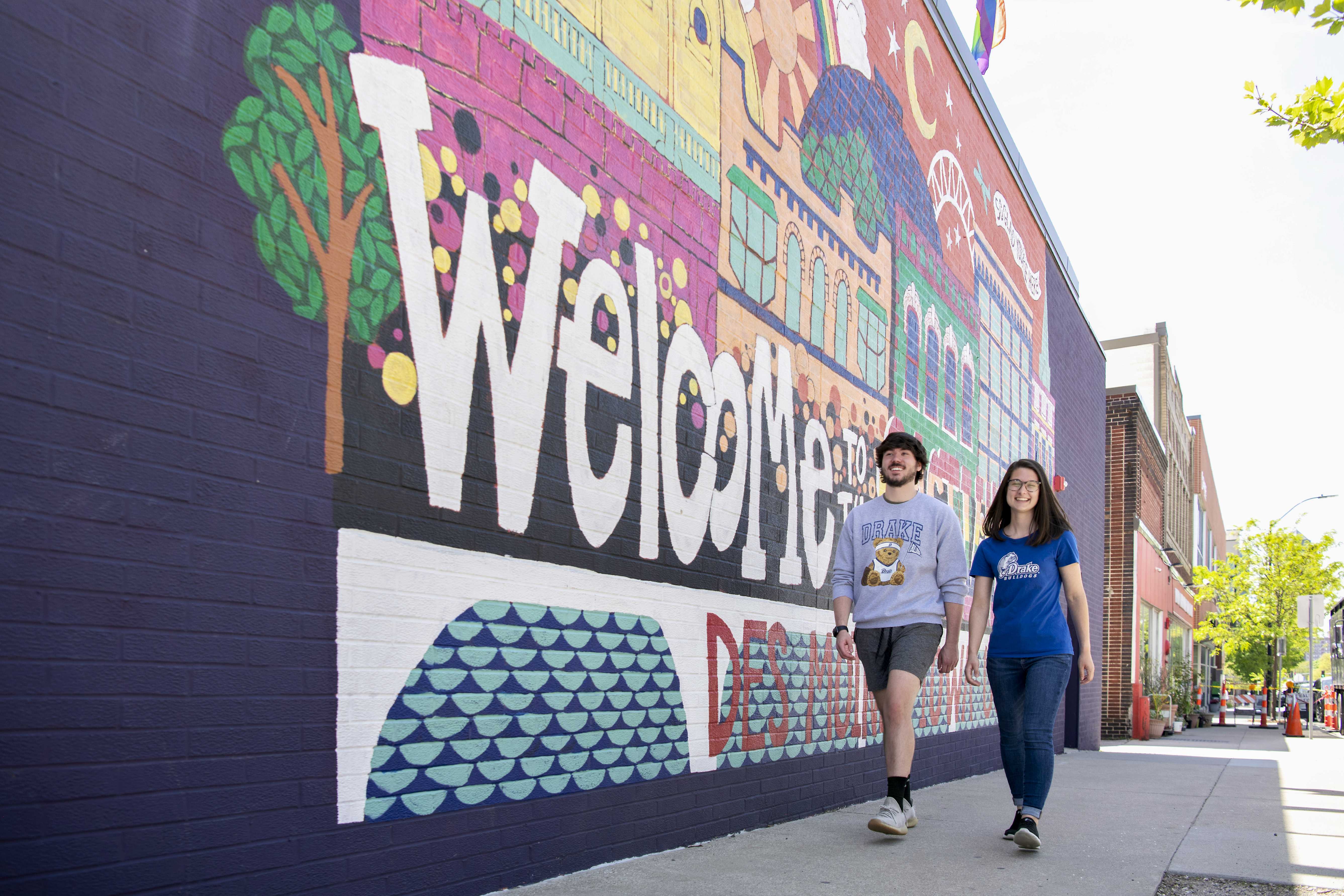 Two students wearing shirts with the Drake logo walk by a colorful mural that reads "Welcome to Des Moines"