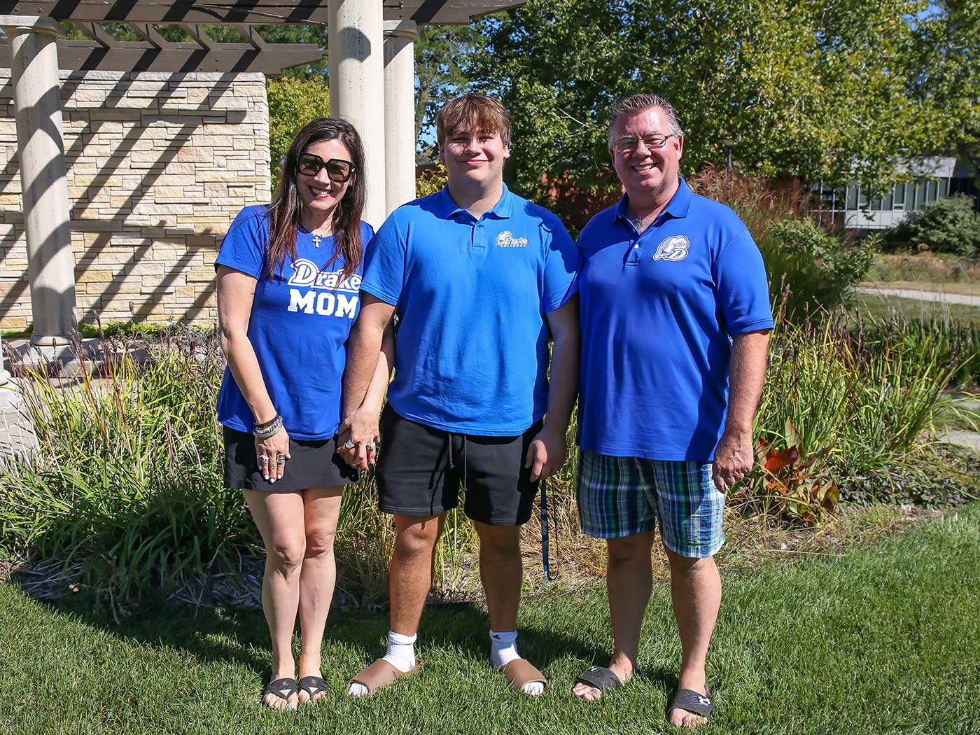Parents and their student, all in blue Drake shirts, smile and stand near an area of campus with tall grass and a stone building