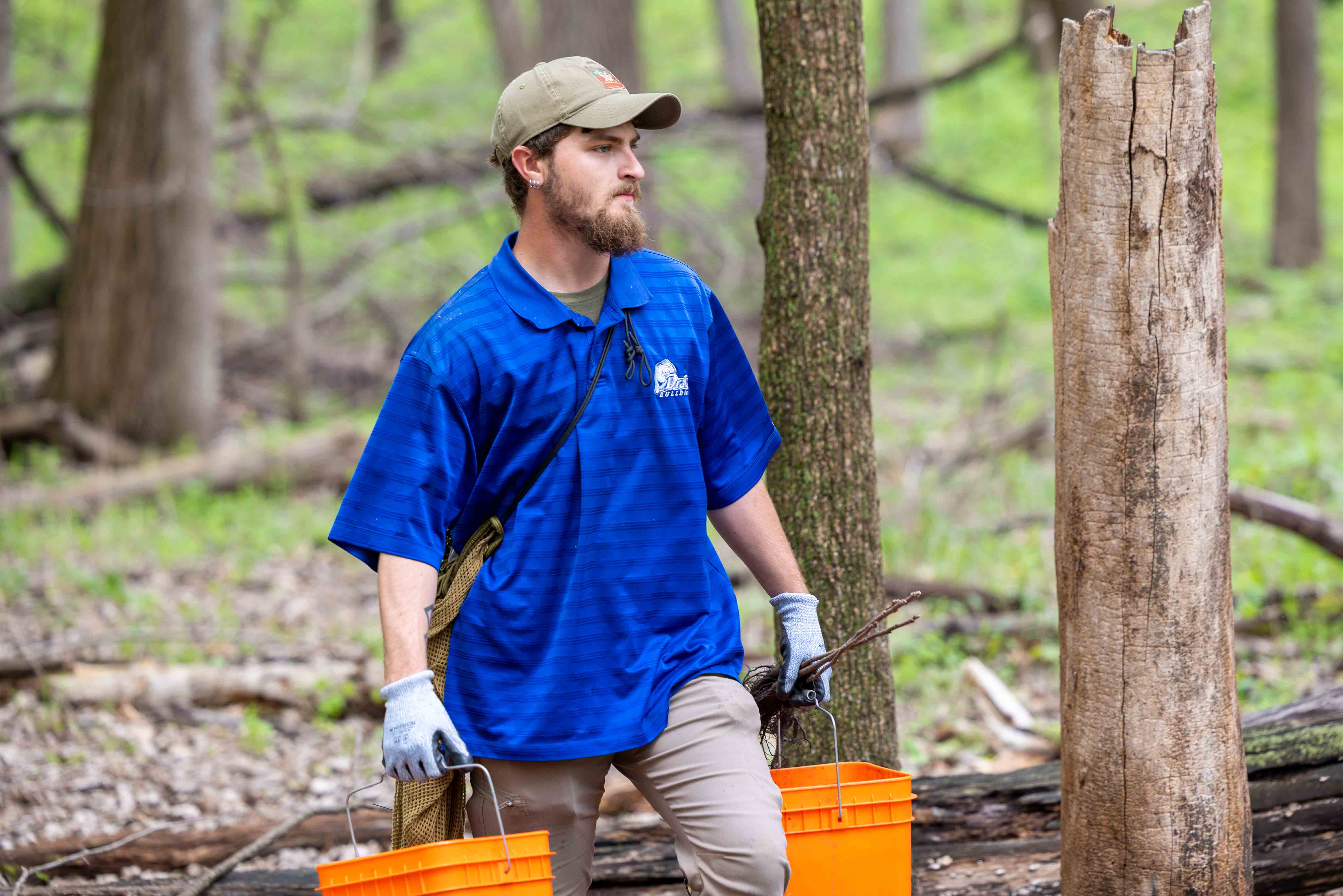 A student wearing a hat and a blue Drake polo carries 2 orange buckets while walking through the woods