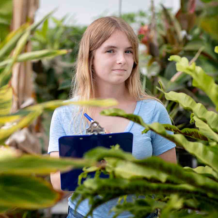 A student smiles at camera, holding a pen and clipboard while standing in a greenhouse