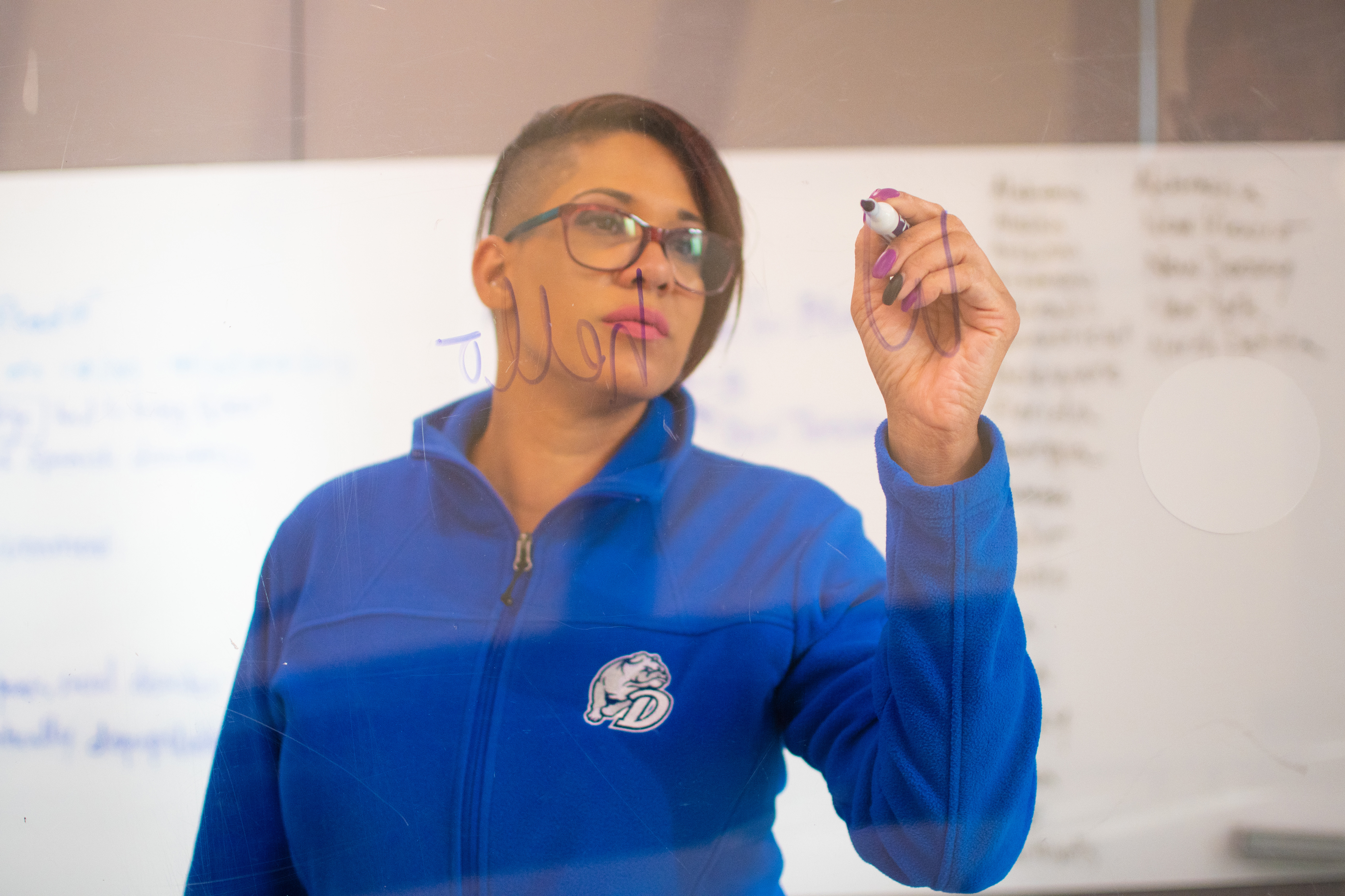 A student in a blue Drake sweatshirt writes with a marker on a clear board