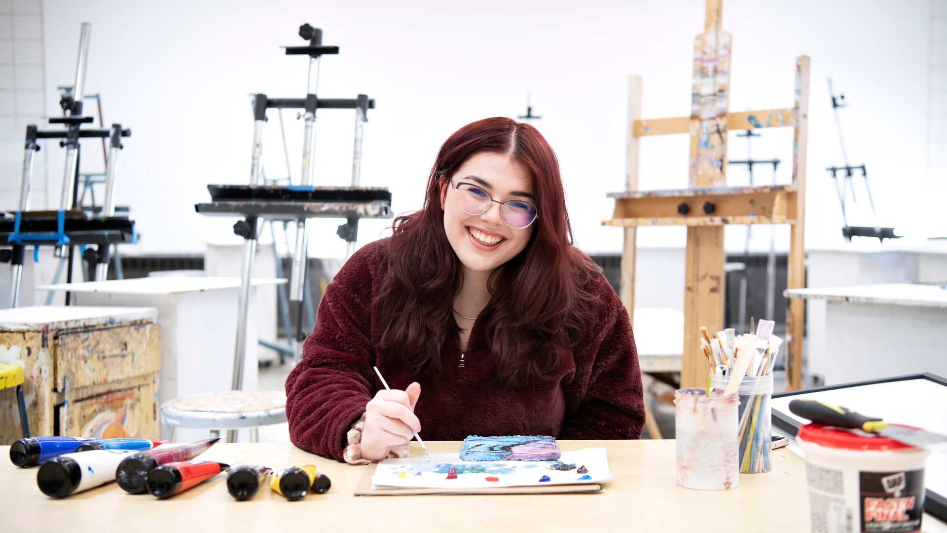 A student smiles at the camera while painting on a canvas on a tabletop in an art studio