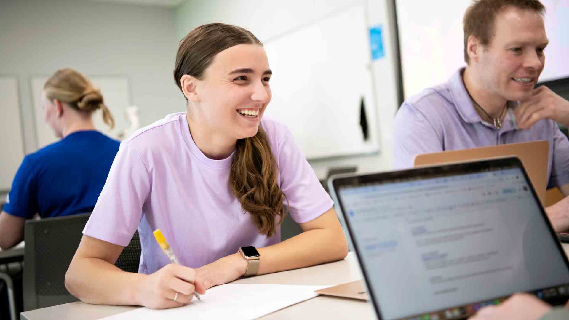 A student smiles at another student across the table while writing in a notebook