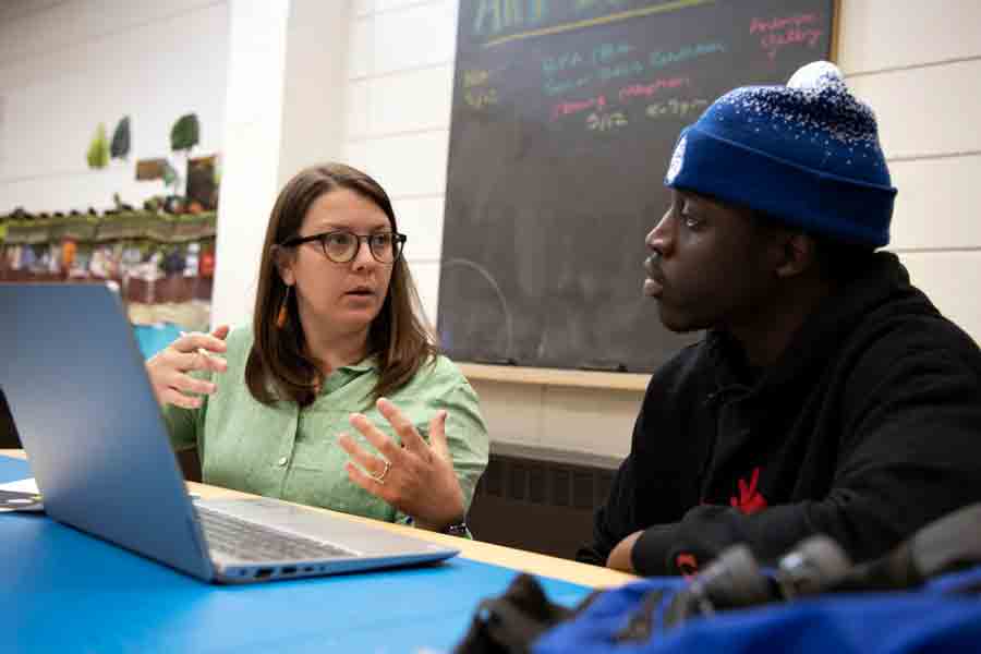 A faculty member speaks to a student while sitting at a table in front of a laptop
