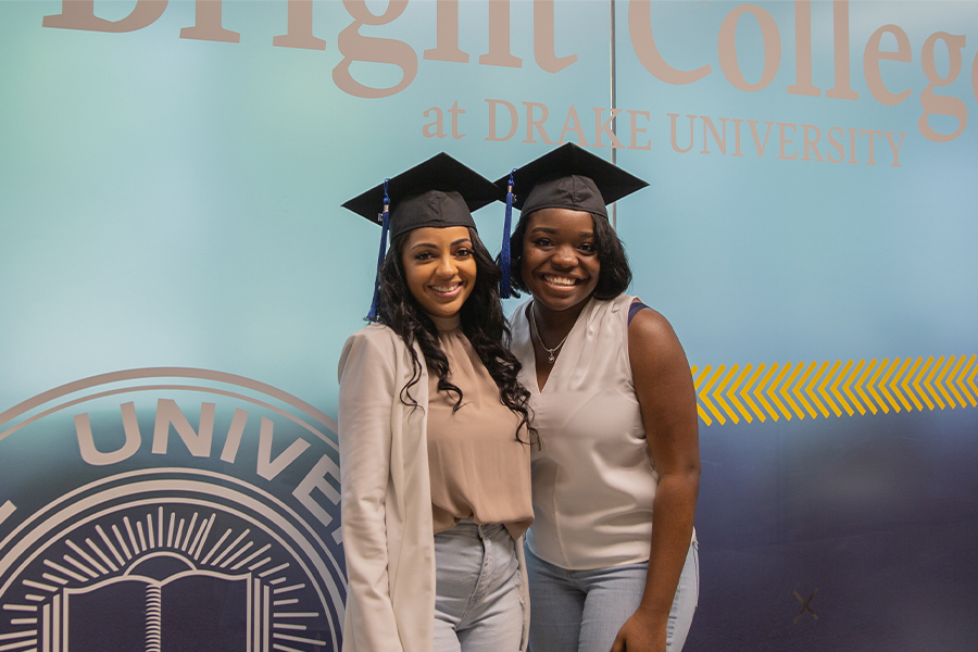 Two students in graduation caps pose in front of the sign for Drake University's Bright College