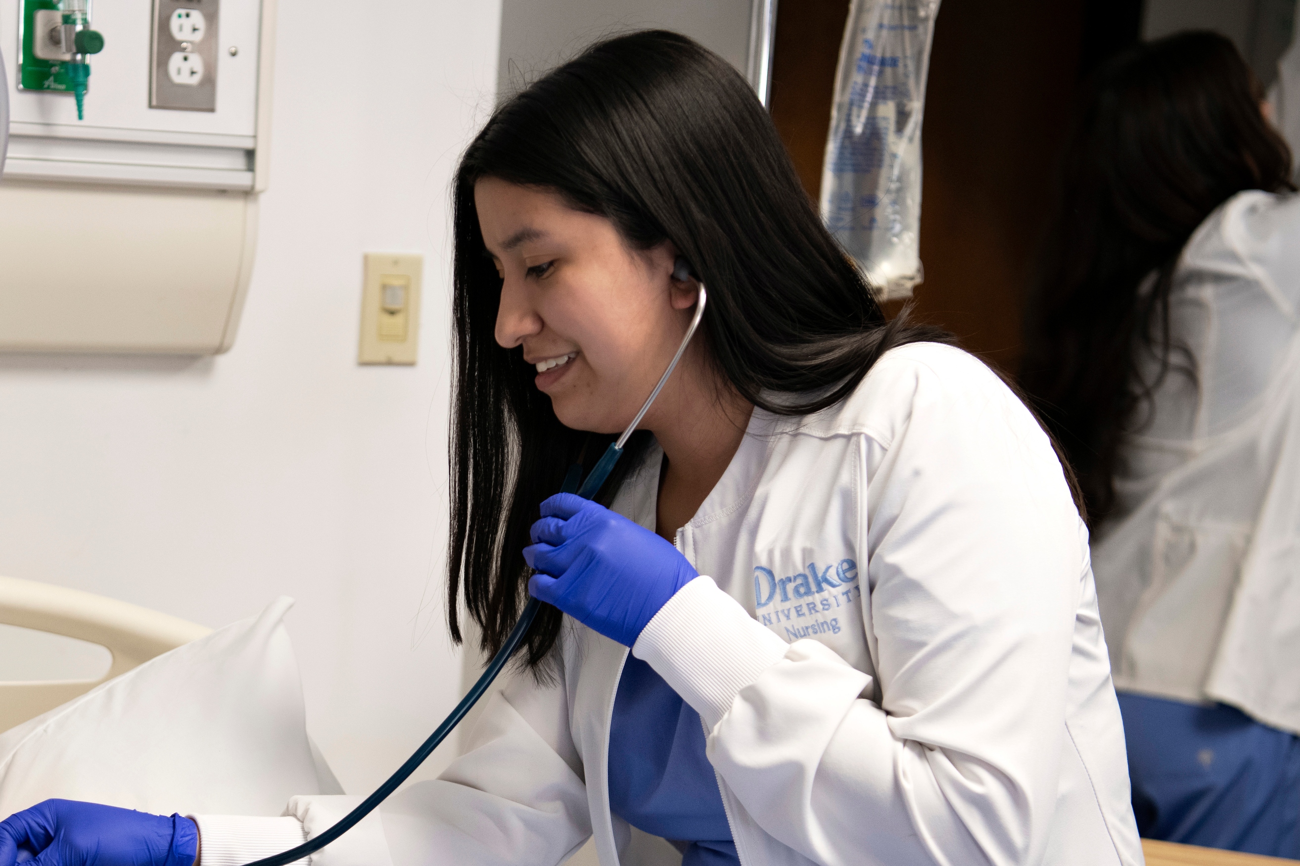 A Drake University student wearing a white coat and blue gloves takes vital signs with a stethoscope during a clinical assignment