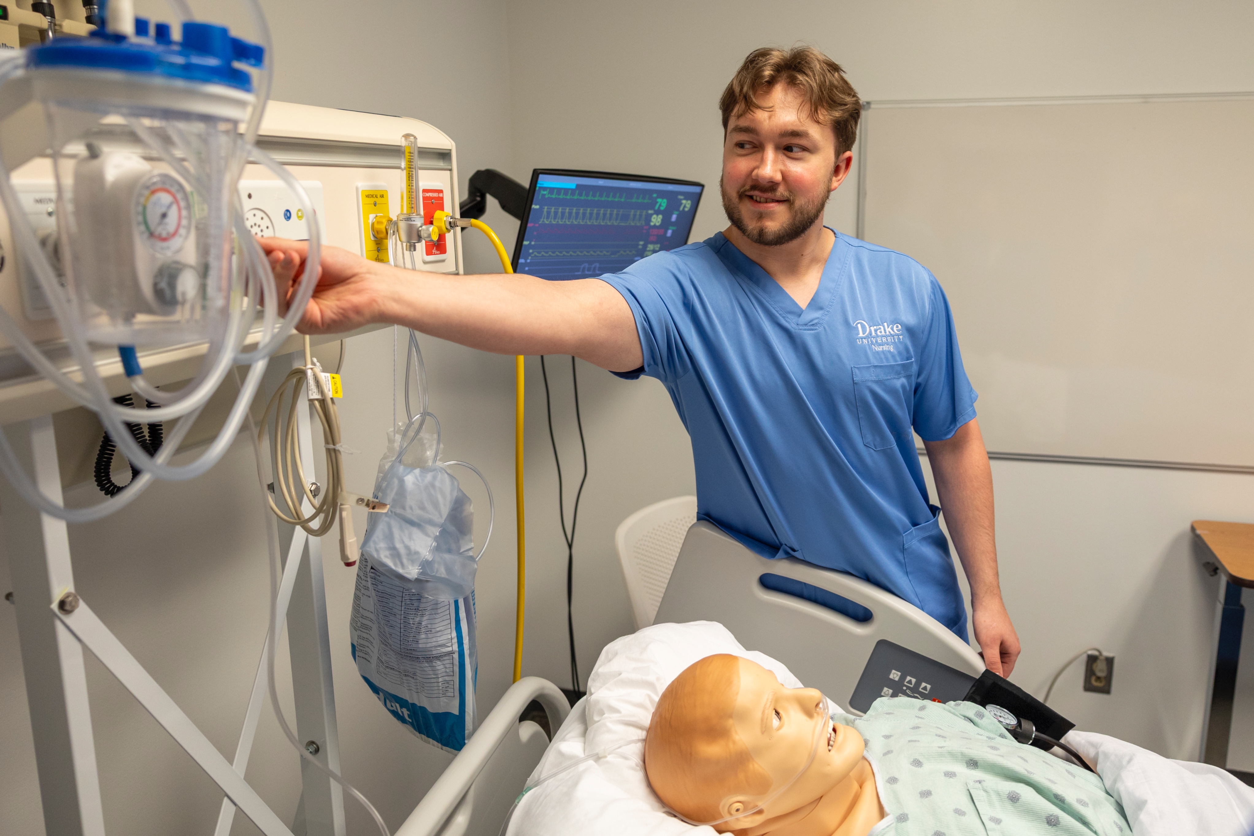 A Drake ABSN student adjusts equipment in a simulation lab where an adult mannequin lays on a bed