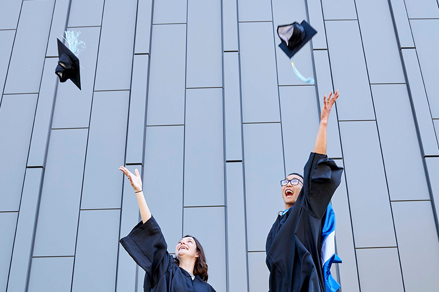 Two students from Drake's Zimpleman College of Business wearing graduation gowns throw their caps up into the air