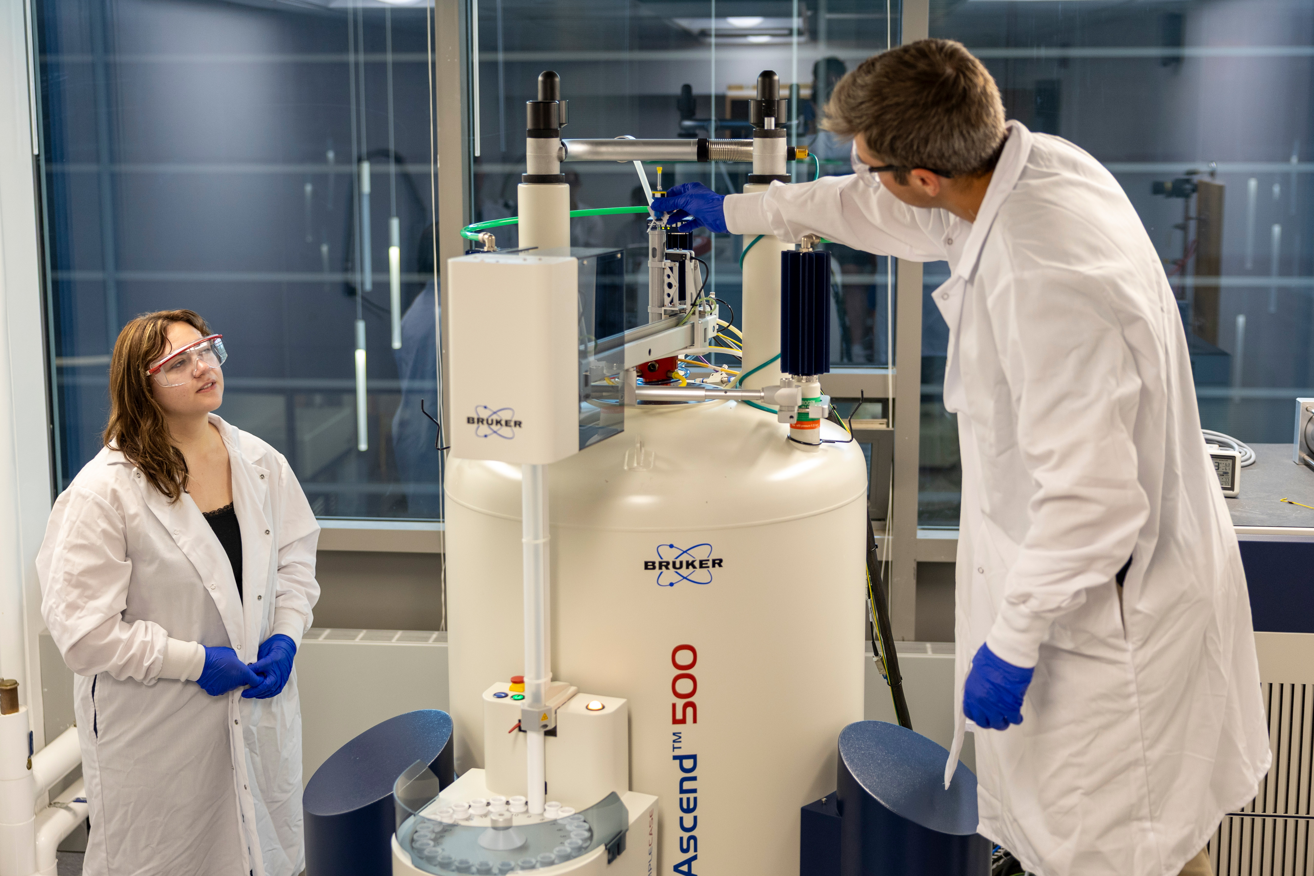 During on off-campus assignment, a Drake Biochemistry, Cell, and Molecular Biology student in a lab coat watches a professional adjust the Bruker Ascend 500 magnet system