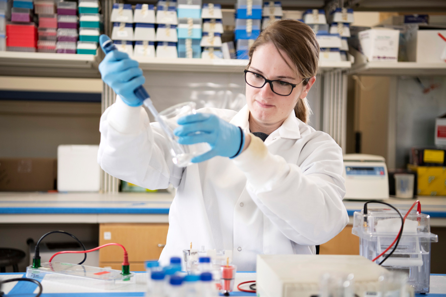 A Drake Biology student in a lab coat and gloves uses a syringe to draw a sample from a beaker