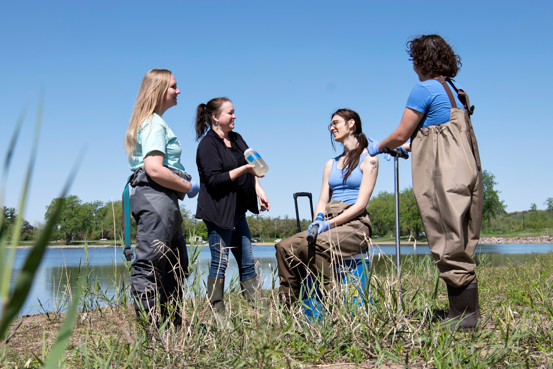 A Drake faculty member explains a water quality sample to three Biology students in waders standing near a pond