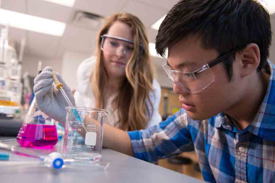 A Drake Chemistry student releases blue liquid from a syringe into an empty graded beaker as a classmate looks on
