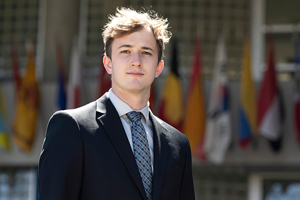 A Drake Data Analytics student in a suit and tie stands outdoors in front of a row of flags