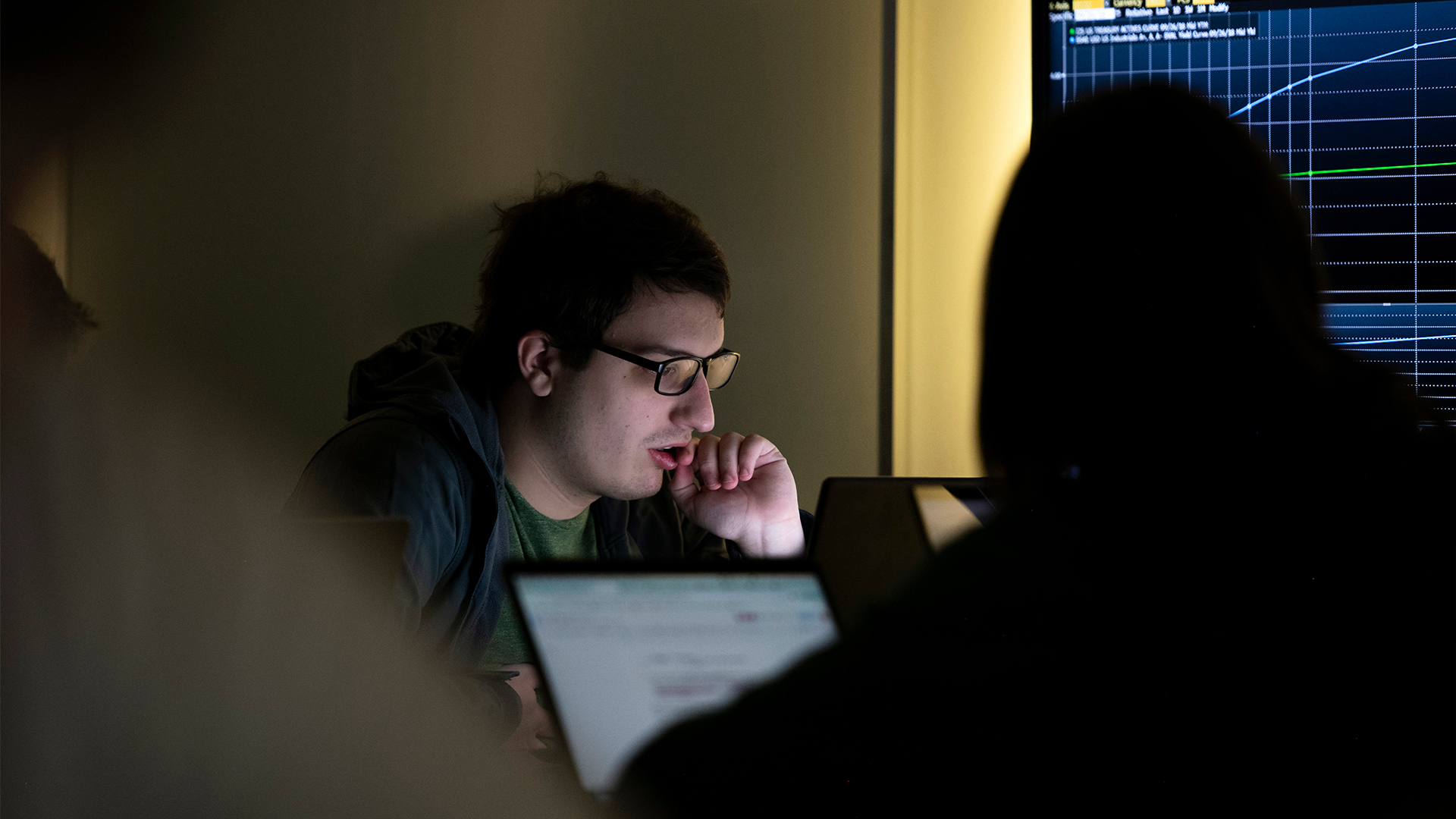 Drake Data Analytics students solve problems in front of laptops and a large monitor in a dark computer lab