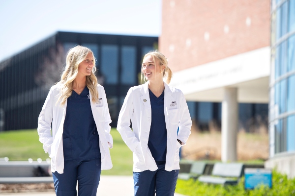 Two Drake PharmD students in white lab coats and blue scrubs walk through campus