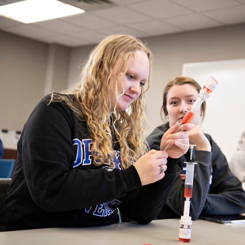 Two Drake students in school sweatshirts practice drawing diabetes medication from a vial in a classroom