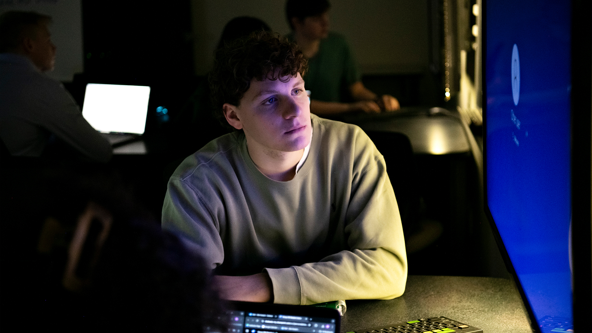 A Drake Economics major studies a large computer monitor in a dark computer lab; behind, other students use their laptops