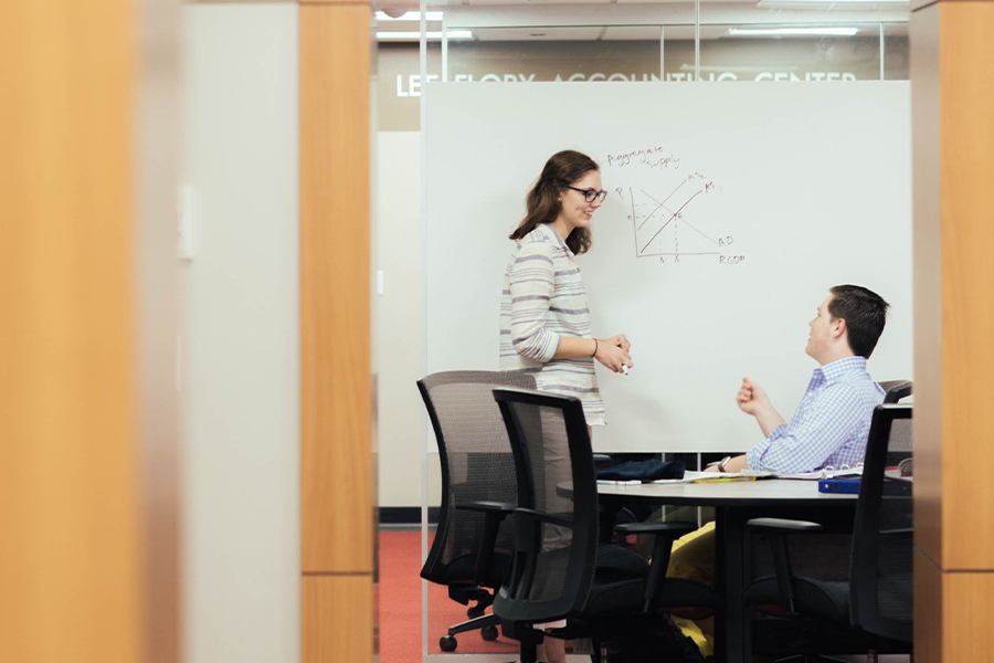 A professor, sitting behind a desk, and an Economics student, standing, discuss a math problem written on a whiteboard