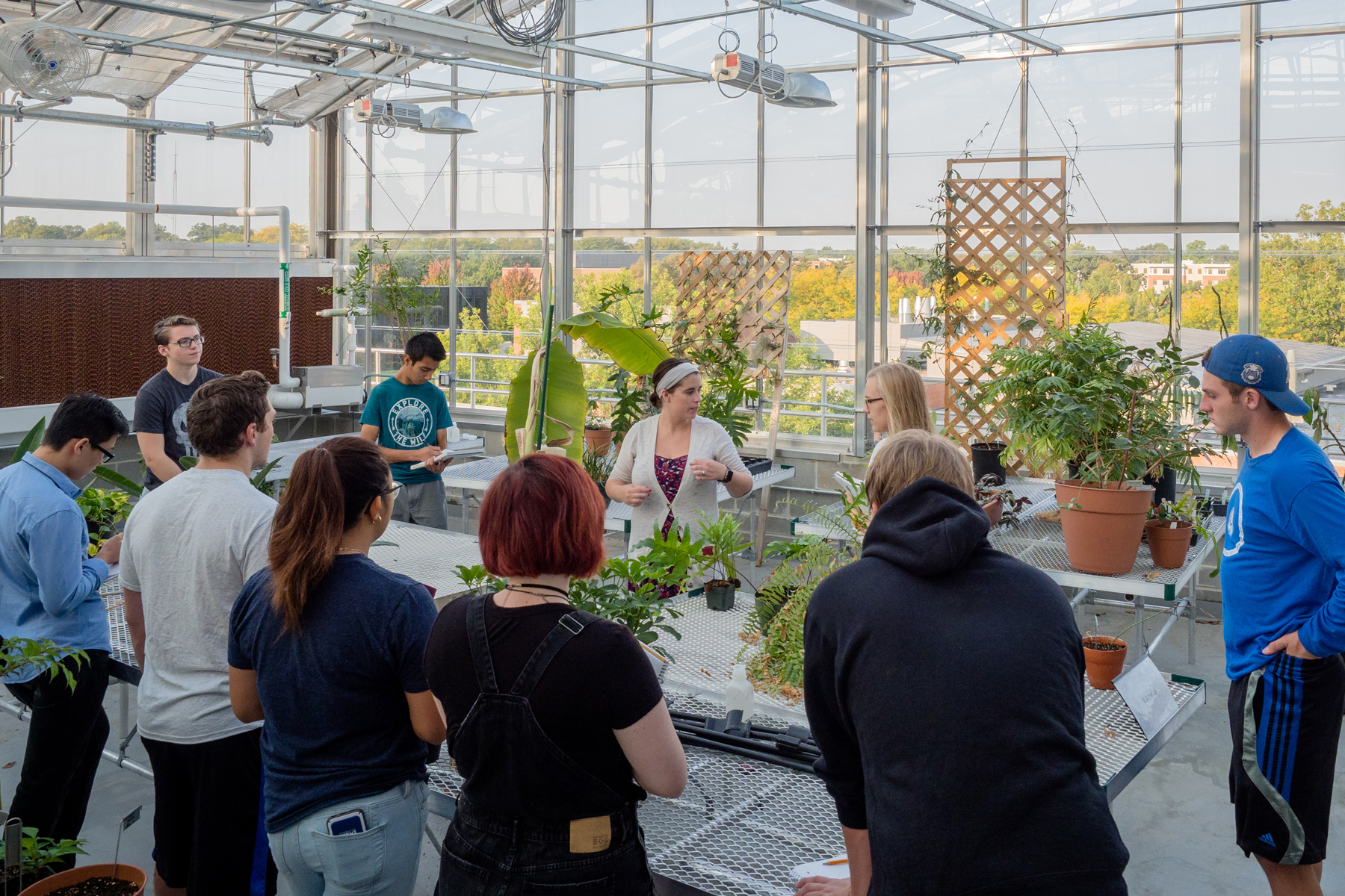An instructor addresses a group of Drake Environmental Science students standing around a table of plants in a greenhouse