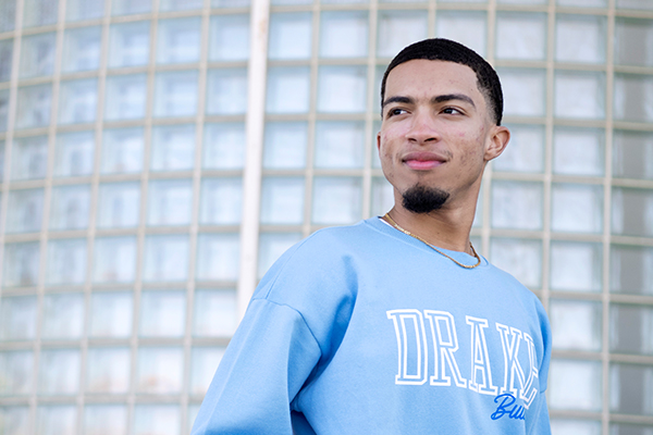 A Drake Finance major in a light blue school sweatshirt stands in front of a glass brick wall
