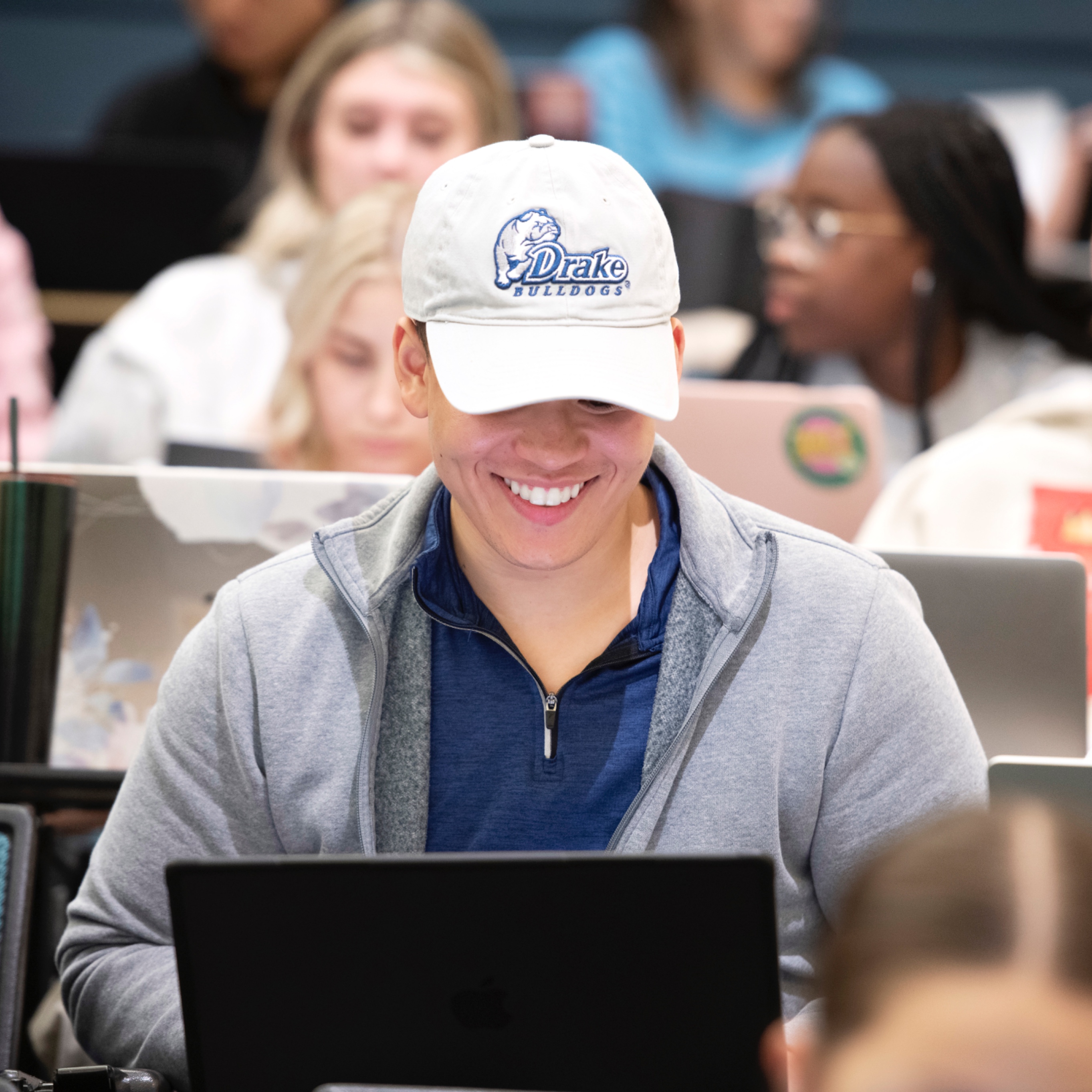 In a class for Drake's Global and Comparative Public Health Concentration, a student wearing a baseball cap smiles as they use a laptop
