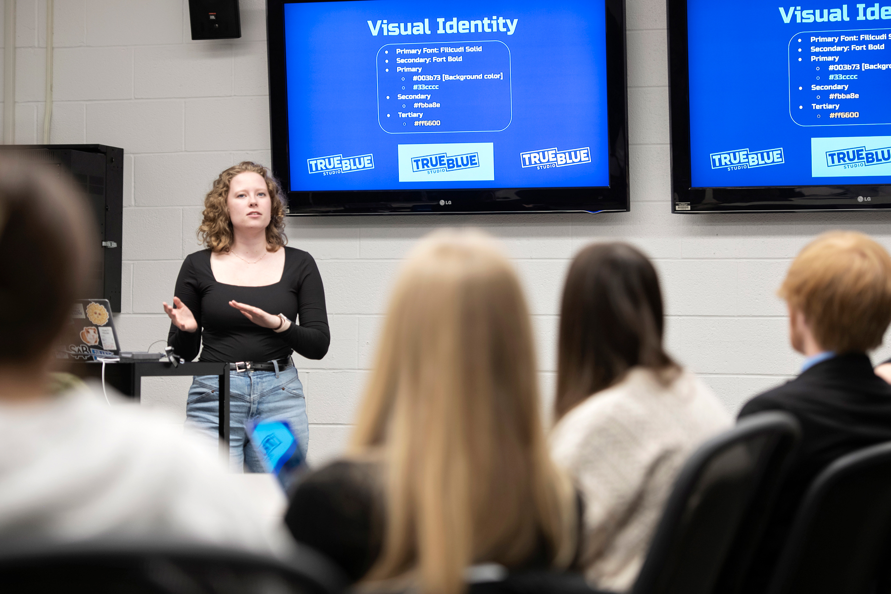 Standing in front of a series of monitors, a Drake Graphic Design student explains their True Blue branding project to a group of classmates