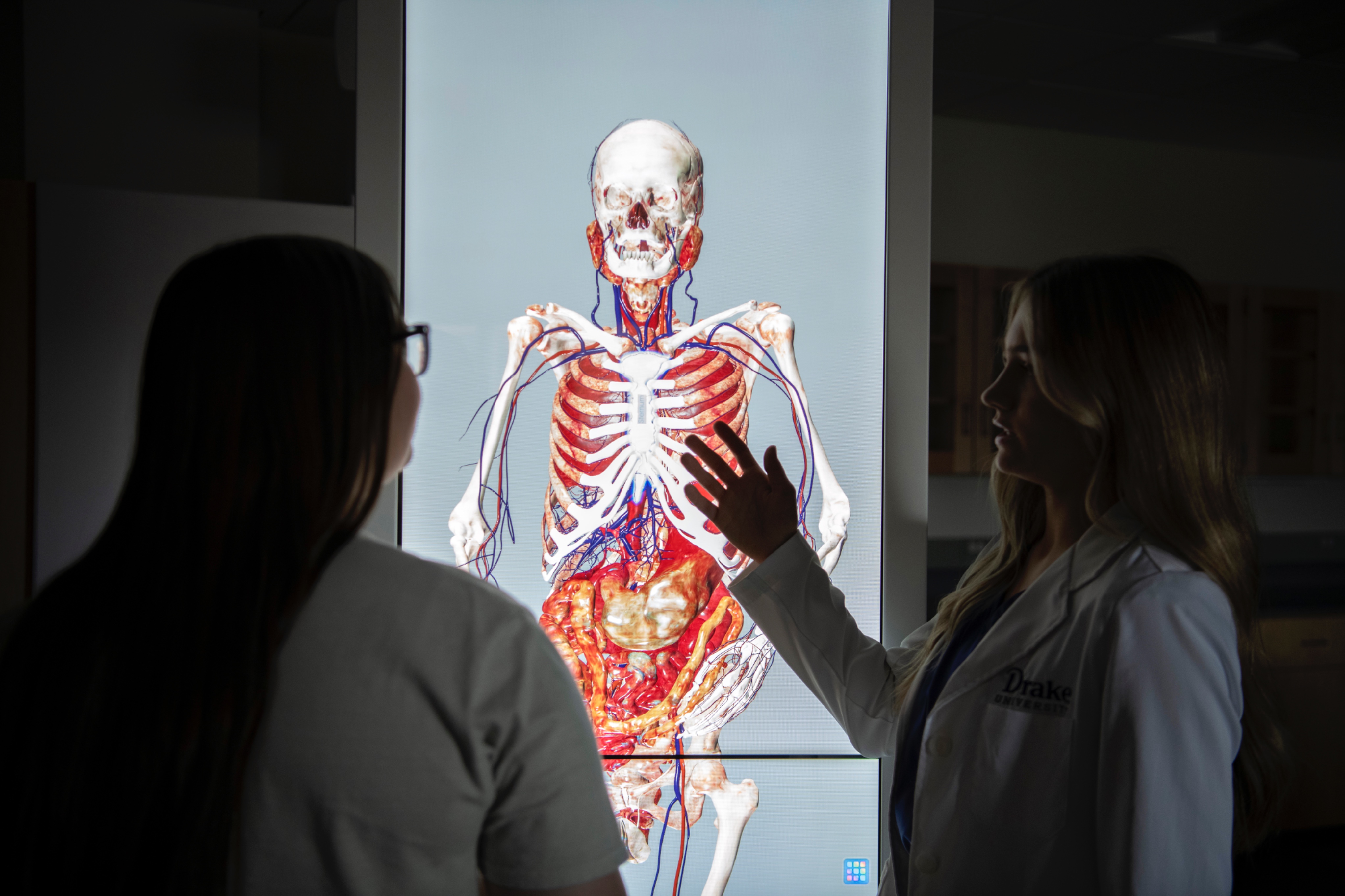 In a dark room, two Drake Health Sciences majors in white coats examine a lit screen with the human body's systems