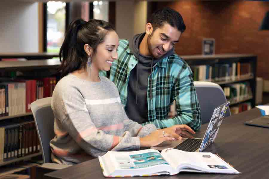 In a library with a textbook and laptop open, two Drake students collaborate on an assignment