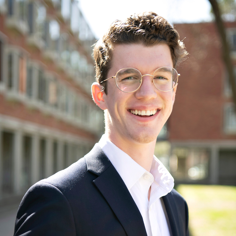 A Drake student in glasses and a suit smiles as they stand outdoors