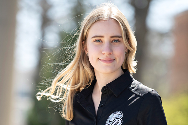 A Drake International Business student, in a black shirt with the Bulldog logo, stands outdoors as the wind blows their hair