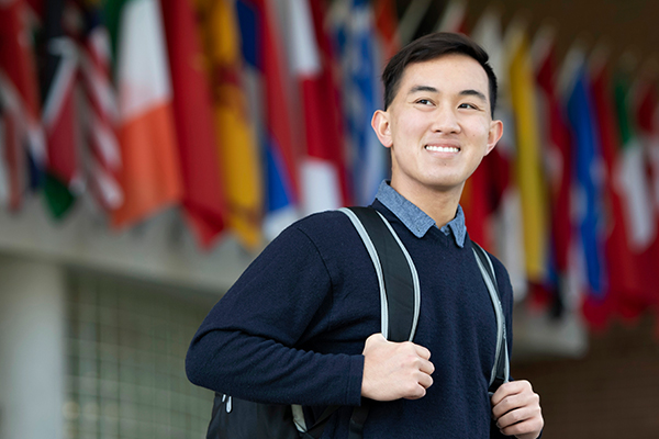 A Drake Law and Business minor loops their fingers through their backpack straps while looking toward the left and standing in front of a line of international flags