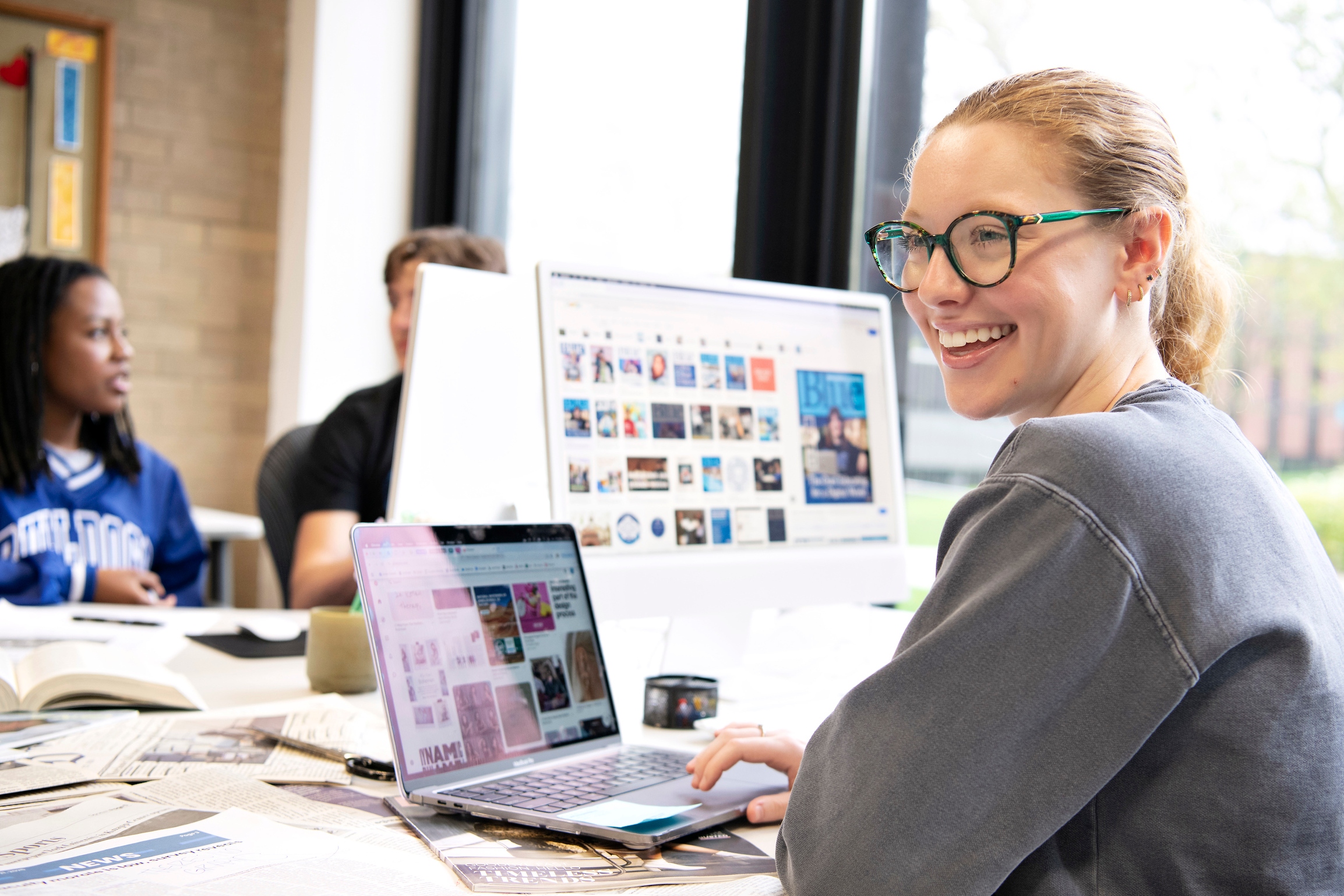 In a campus computer lab, a student from Drake's Magazine and Brand Media program examines a series of images on a laptop screen while sitting near a larger monitor