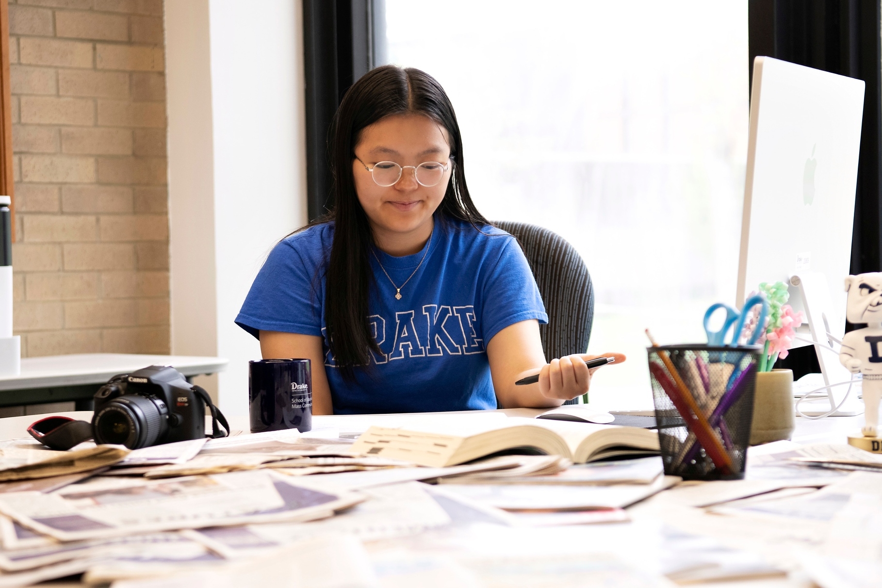 A student in Drake's Magazine and Brand Media program reviews a large textbook in a campus media lab, with a camera and coffee mug situated on the table top