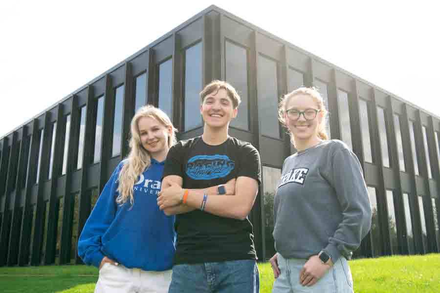 Three journalism students from Drake smile while standing in front of a black building with long windows