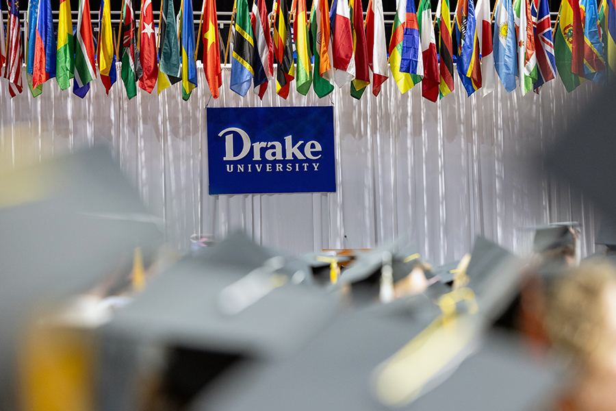 Signage for Drake University situated below a series of international flags and behind a group of students in graduation caps
