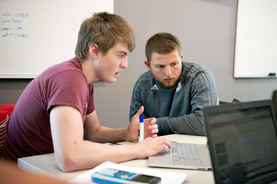 In a classroom, one student working at a laptop explains a concept to a Mathematics for Secondary Education major during a tutoring session