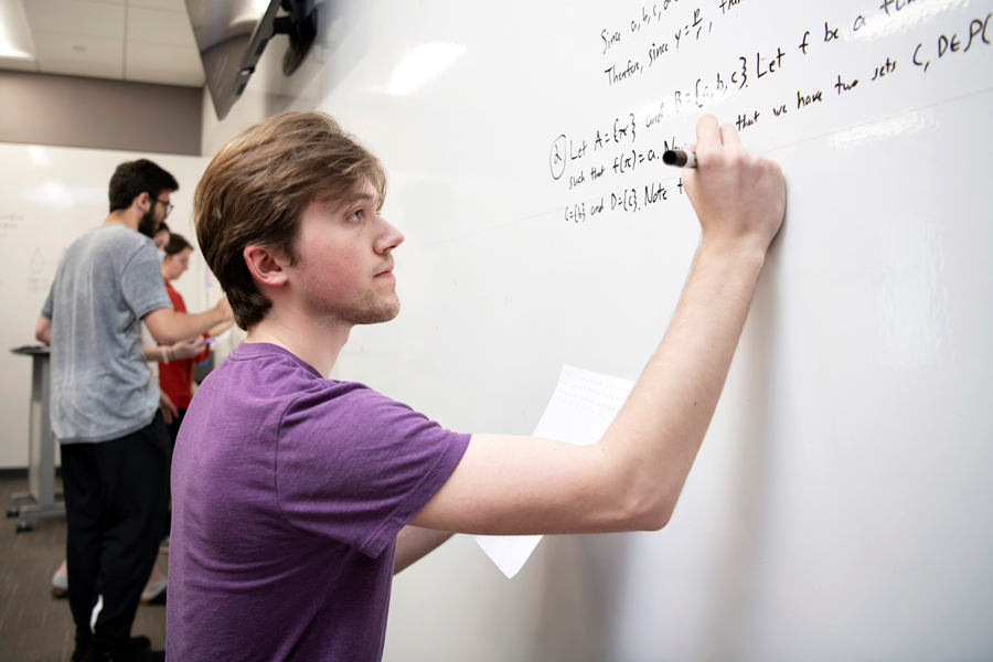 In a classroom lined with whiteboards, a student in Drake's Mathematics for Secondary Education program writes a lengthy equation and their logic with a black marker