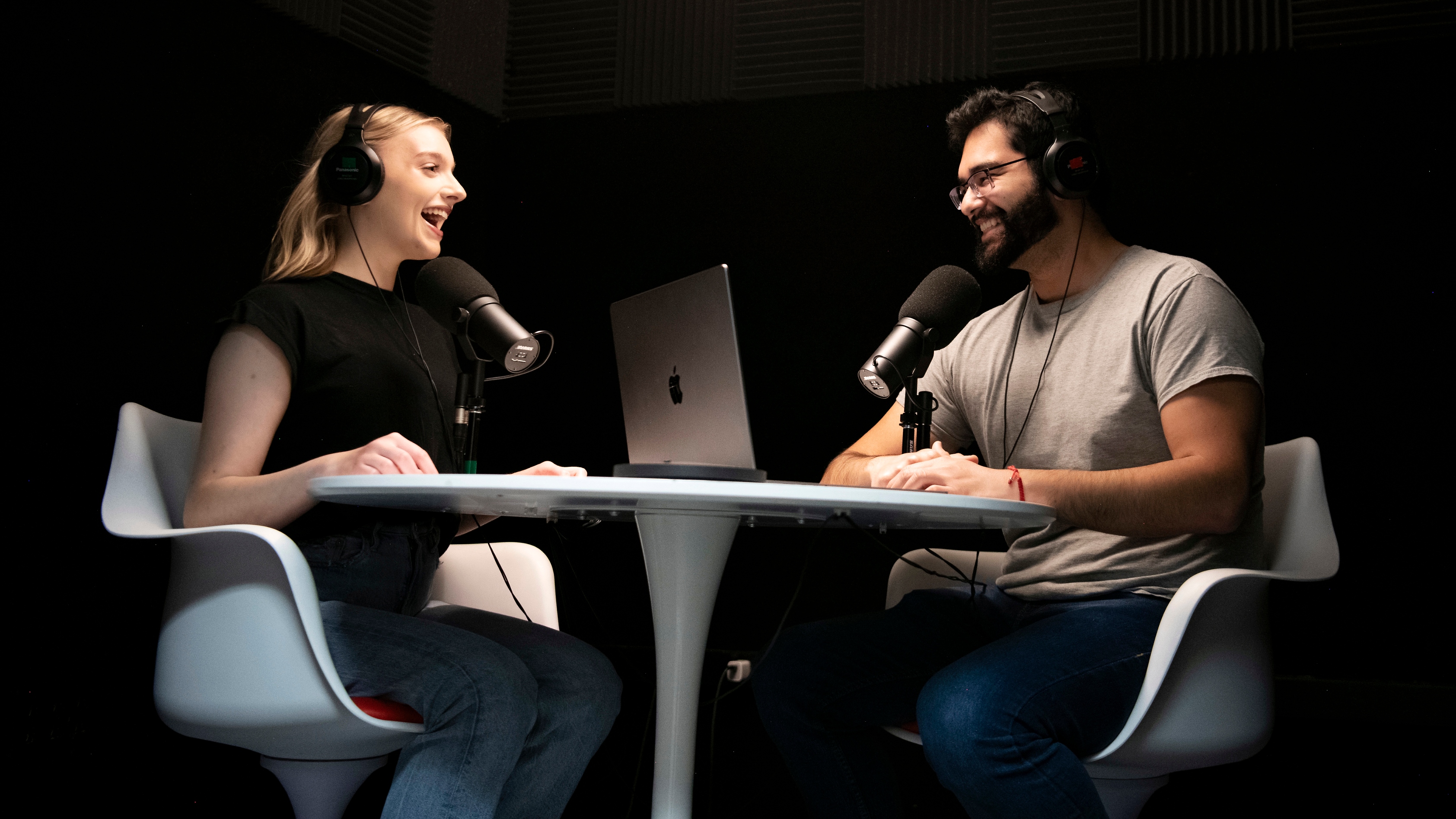 Two Drake Multimedia Journalism Majors conduct a podcast with laptops, microphones, and headphones around a white table
